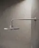 Duschkopfanschluss von Ideal Standard, montiert an einer Wand aus hellem, strukturiertem Material. Schwenkbarer Arm mit flacher, runder Kopfplatte.